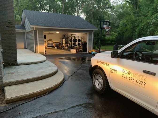 A landscaping company truck is parked on a driveway outside a well-lit garage, with gardening equipment visible inside and wet pavement suggesting recent rain or cleaning.