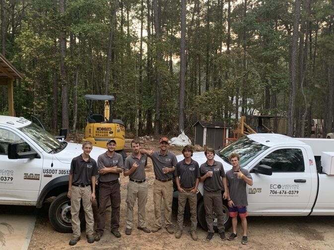 Seven people in uniform stand in front of two work trucks and a small excavator, surrounded by trees.