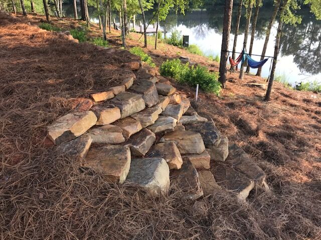 Stone steps lead down a pine needle-covered hill towards a lake, surrounded by trees. Two hammocks are hung in the background near the water.