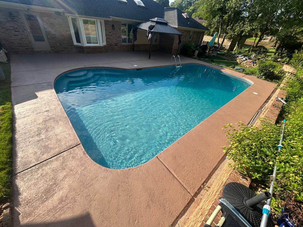 A clear swimming pool with a concrete deck in the backyard of a brick house, surrounded by green grass and plants, under a sunny sky.