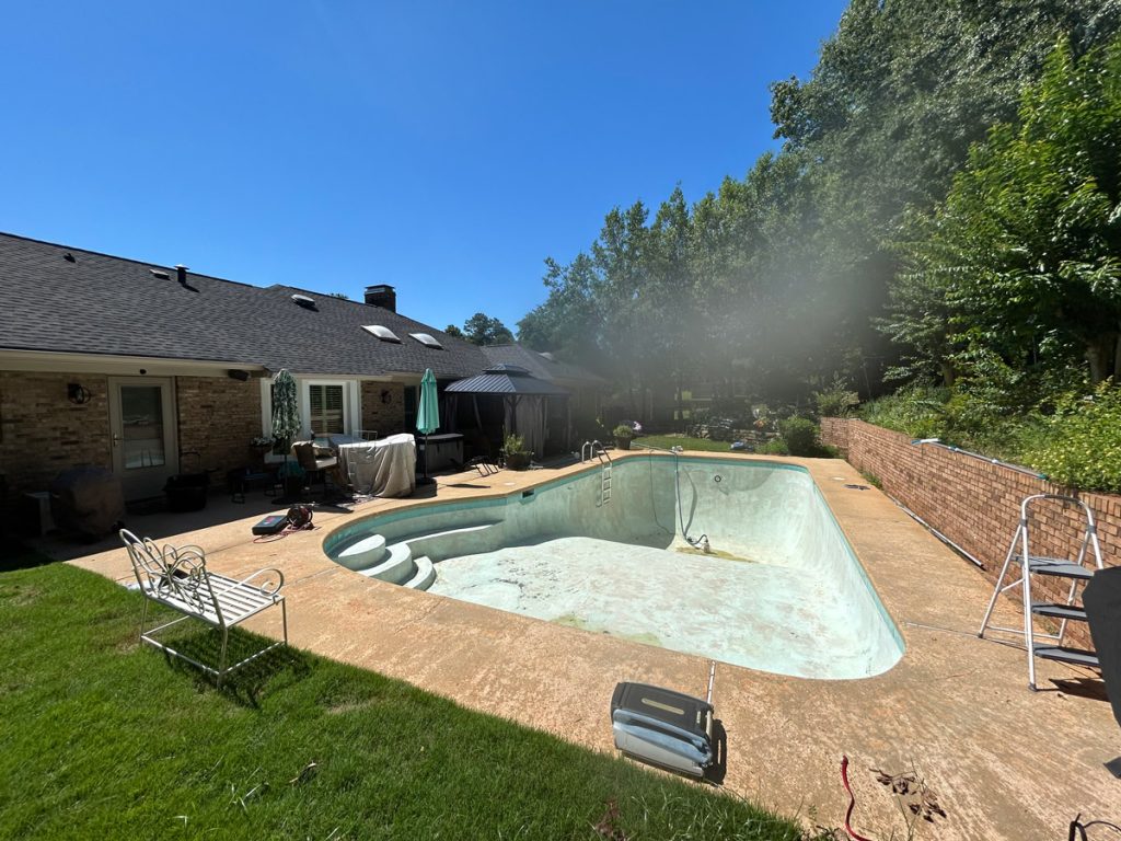 An empty backyard swimming pool with cleaning equipment around it, next to a brick house and patio furniture under clear blue sky.