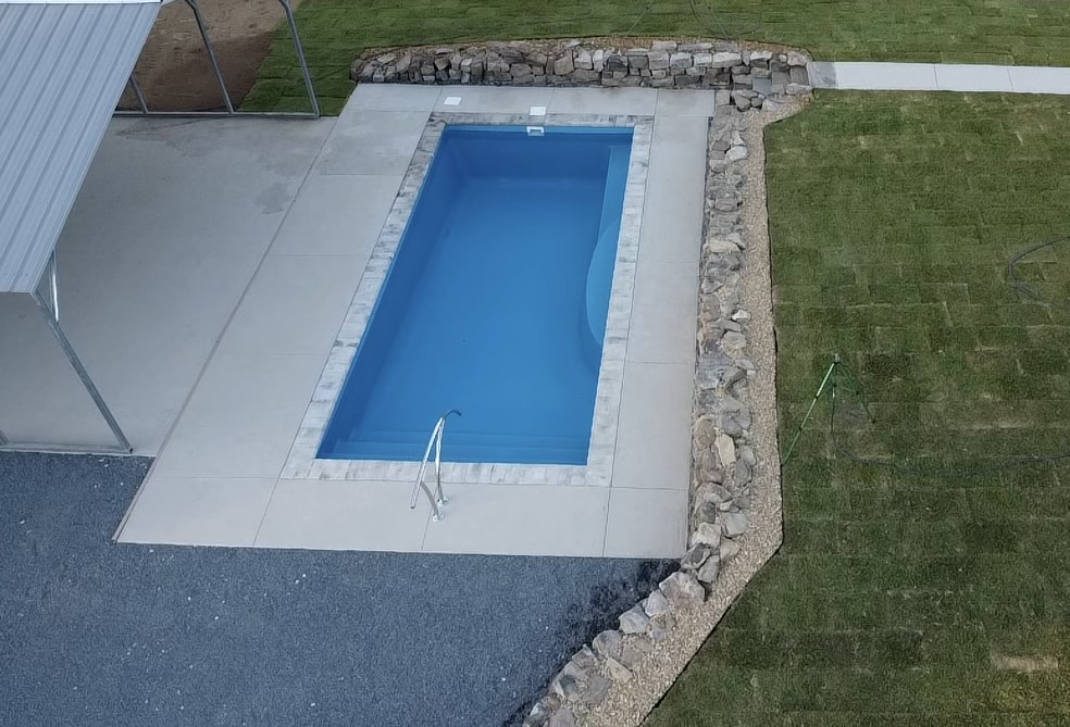 Aerial view of a rectangular pool surrounded by gray concrete and stone edging, adjacent to a grassy lawn.