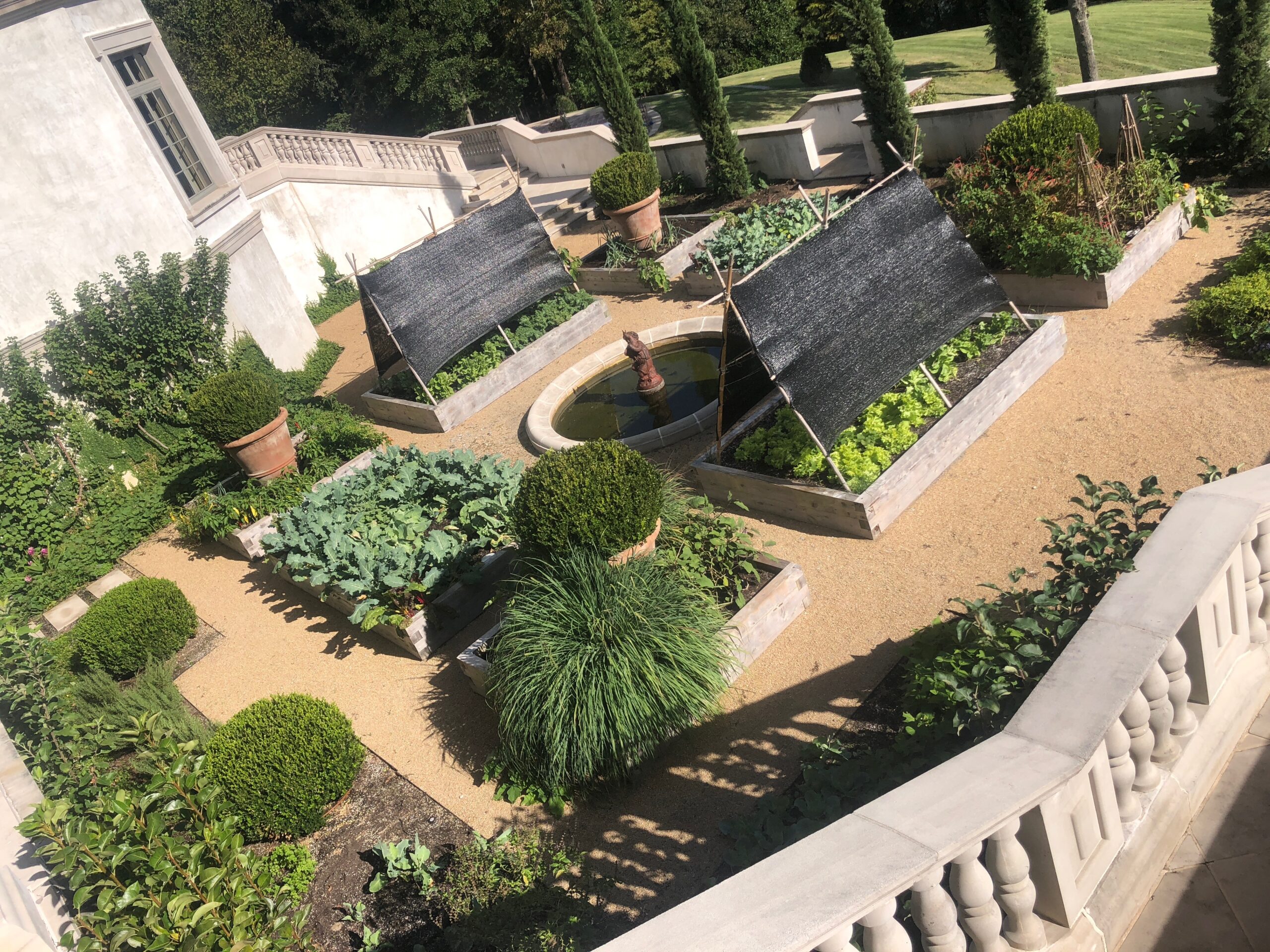 A formal garden with raised vegetable beds, a small fountain, and round topiary bushes, surrounded by a gravel path and stone balustrade.