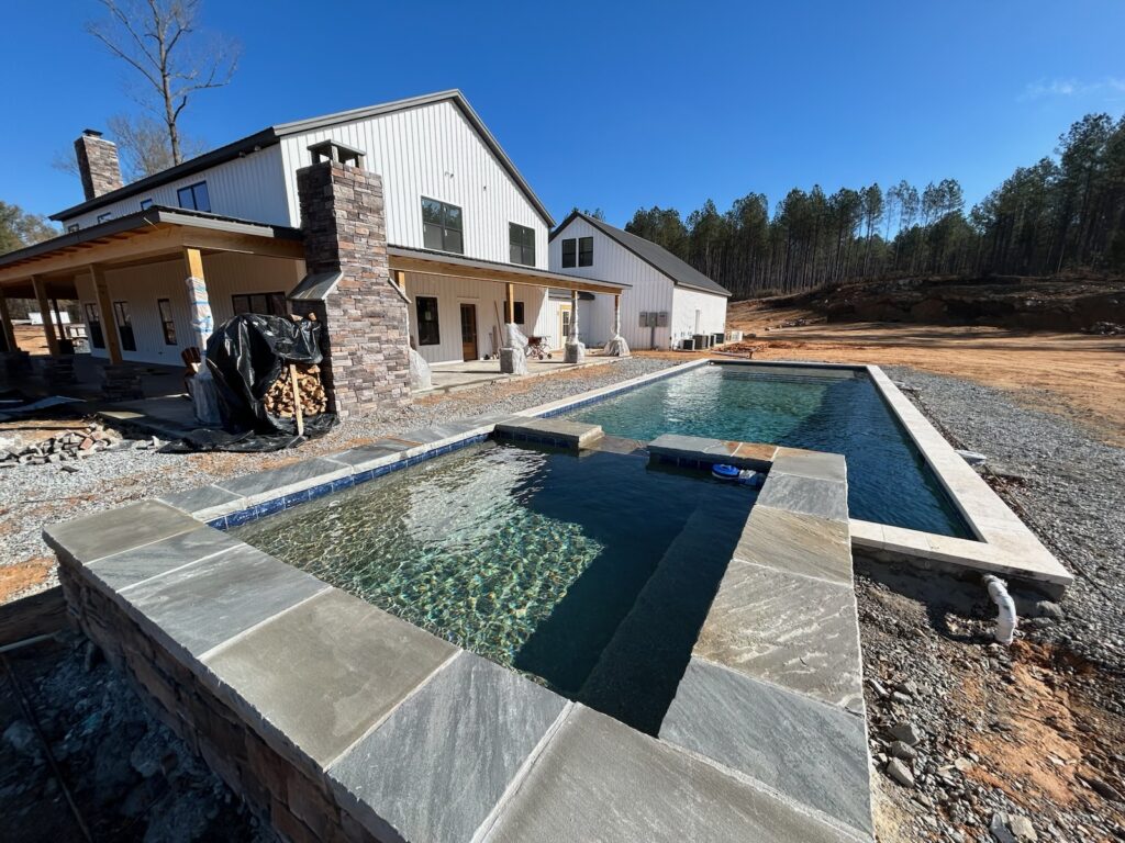 A modern farmhouse with a large covered patio, stone fireplace, and a rectangular swimming pool and hot tub in a partially landscaped yard.