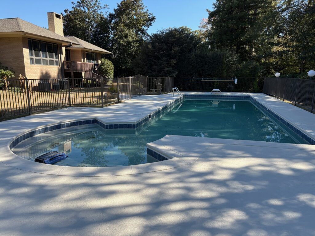 A backyard with an in-ground swimming pool and adjacent hot tub, surrounded by a black metal fence, near a brick house and trees.