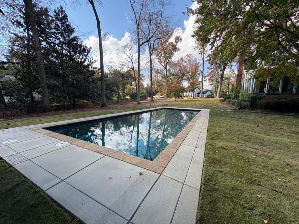 Rectangular outdoor swimming pool surrounded by concrete decking in a grassy backyard with trees and houses in the background.