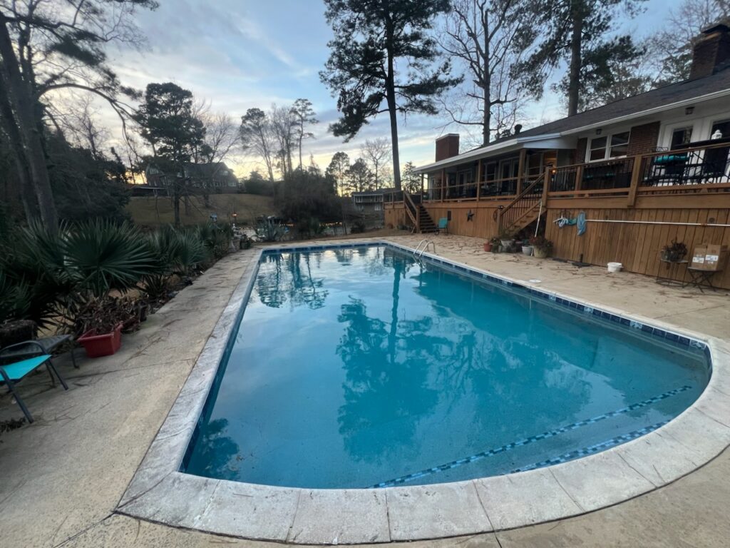 Rectangular outdoor swimming pool next to a wooden deck and house, surrounded by trees and plants under a cloudy sky.
