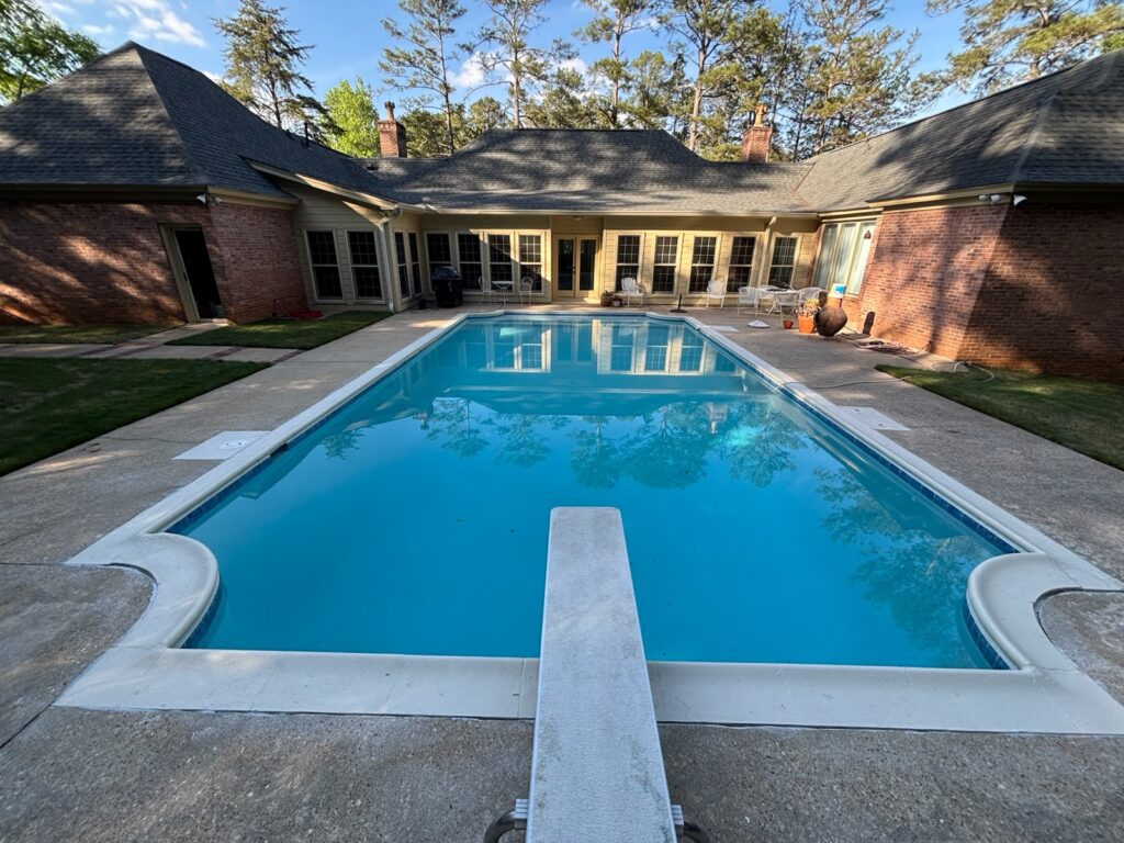 A rectangular outdoor swimming pool with a diving board is surrounded by a concrete deck, adjacent to a brick house with large windows and patio furniture.