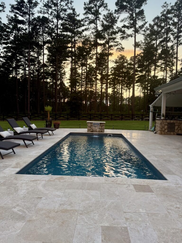 A rectangular outdoor pool with lounge chairs on one side, surrounded by a tiled deck; tall pine trees and a covered patio are visible in the background at sunset.