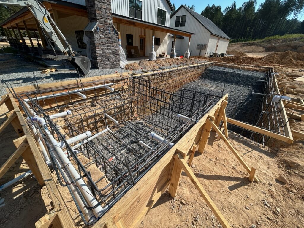 A rectangular in-ground swimming pool under construction with exposed rebar, plumbing pipes, and wooden framing next to a house.