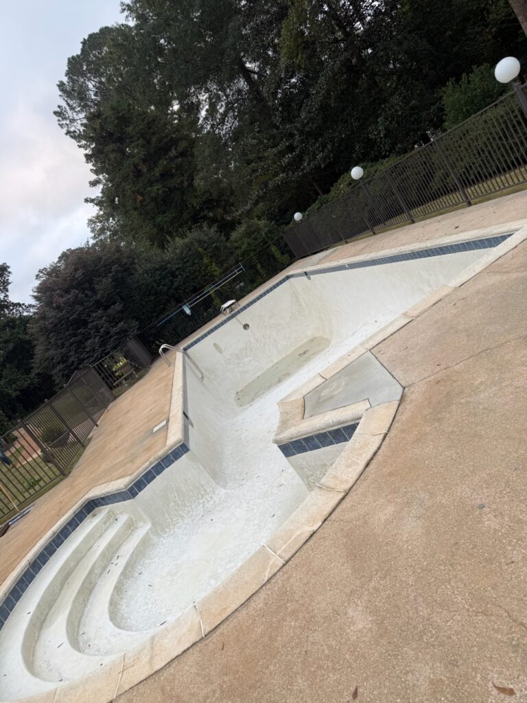 An empty outdoor swimming pool with steps, surrounded by a concrete deck and a black metal fence, with trees and lamp posts in the background.