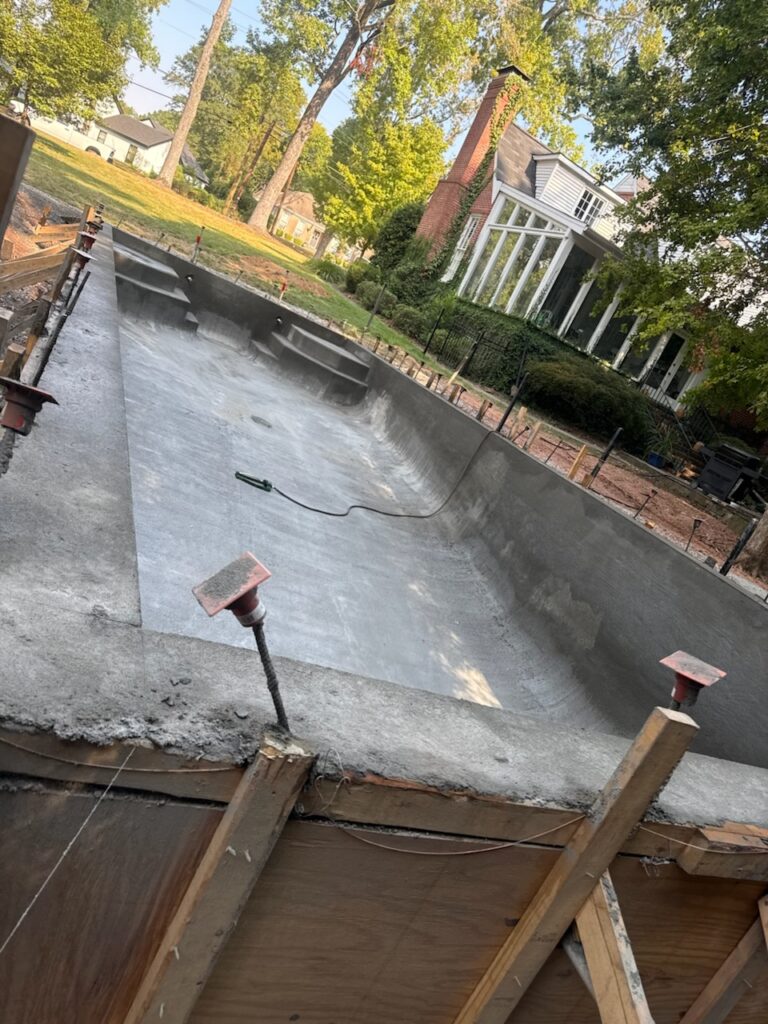 A concrete in-ground swimming pool under construction in a residential backyard, with surrounding wooden forms and tools visible.