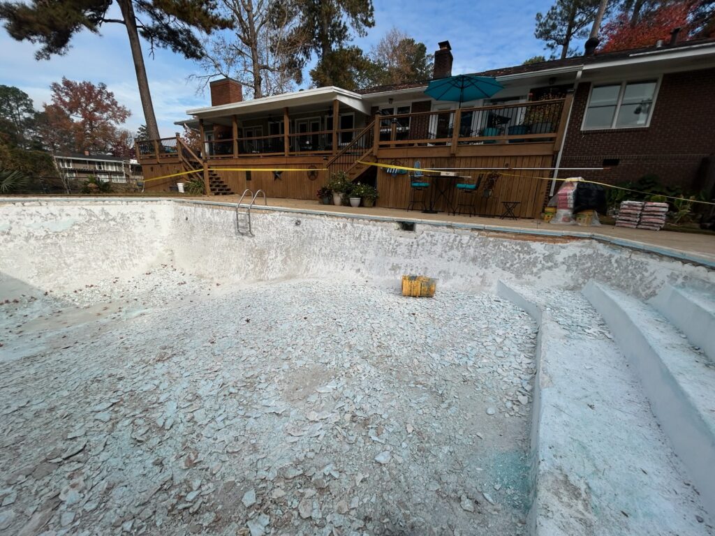 An empty, deteriorating swimming pool with peeling paint sits in a backyard; a yellow bucket and construction materials are visible nearby, and a house with a deck is in the background.