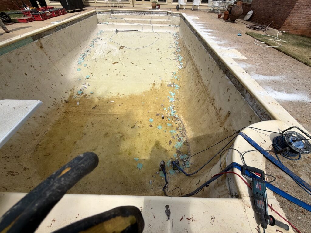 Empty swimming pool with dirty, stained surfaces and broken blue tiles scattered on the bottom. Tools, cables, and cleaning equipment are visible around the pool's edge.