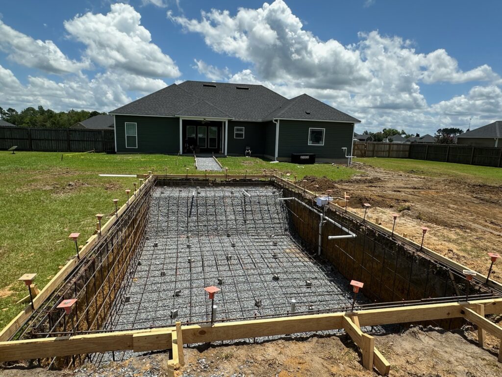 Rectangular backyard swimming pool under construction, showing rebar framework and wooden framing, with a house and cloudy sky in the background.