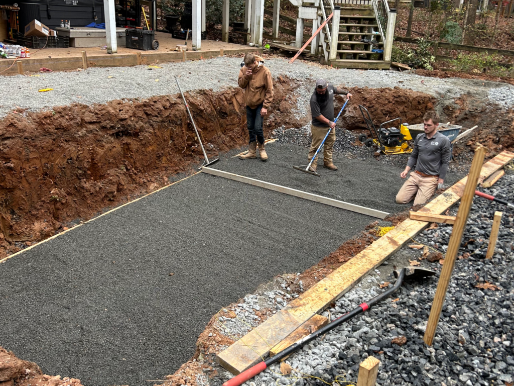 Three people work on leveling gravel in a rectangular foundation pit, surrounded by wooden boards and tools.