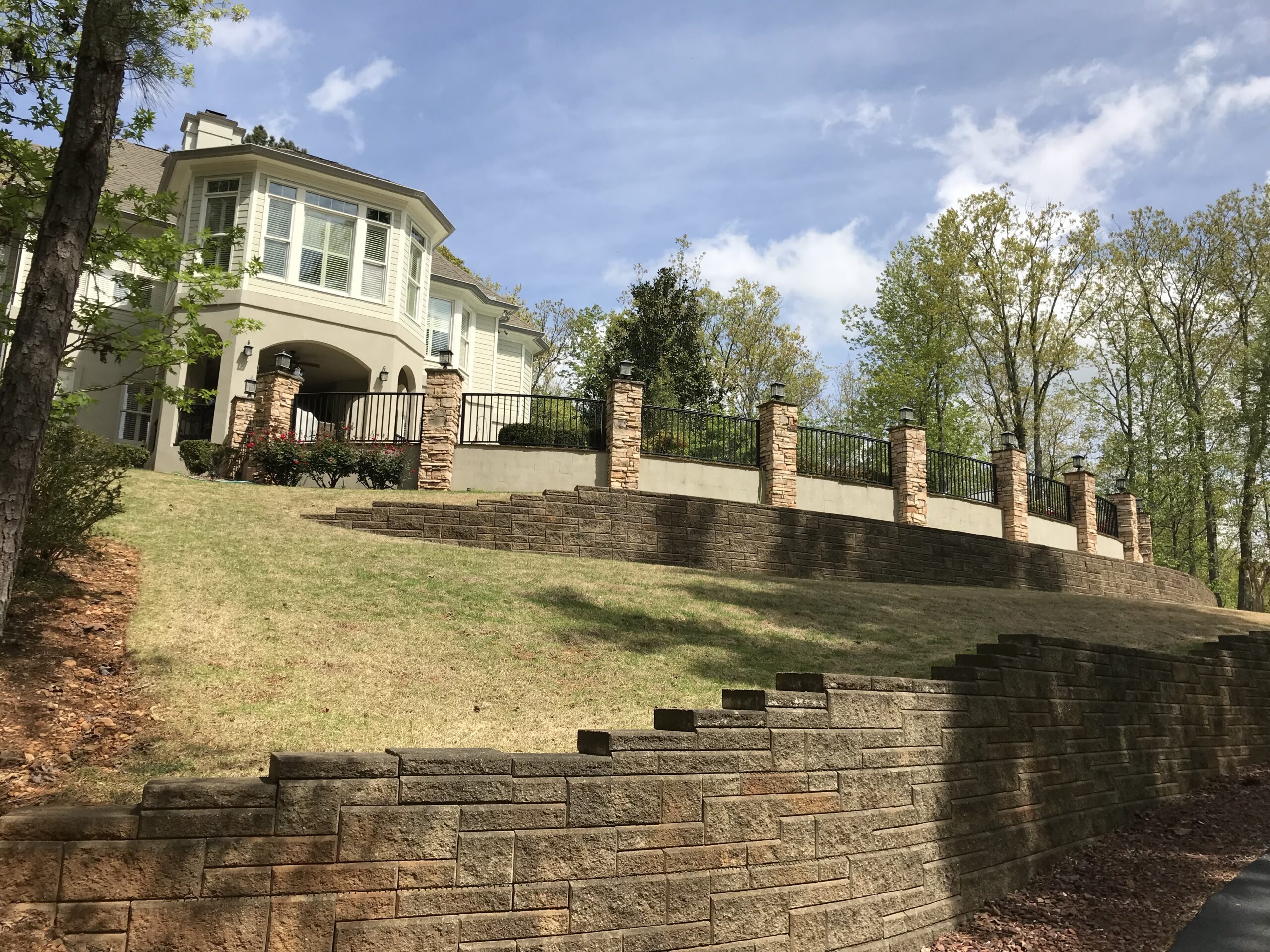 A large white house with a stone fence sits atop a sloped lawn, enhanced by expertly designed hardscaping retaining walls and surrounded by trees under a partly cloudy sky.