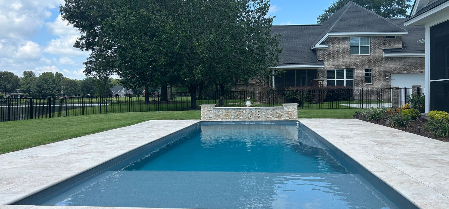 Rectangular backyard swimming pool with light stone deck, small fountain feature, and a brick house in the background surrounded by trees and a black metal fence.