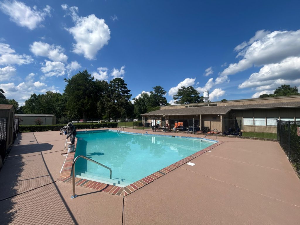 Outdoor swimming pool with metal handrails, surrounded by a concrete deck, lounge chairs, and a building in the background under a partly cloudy sky.