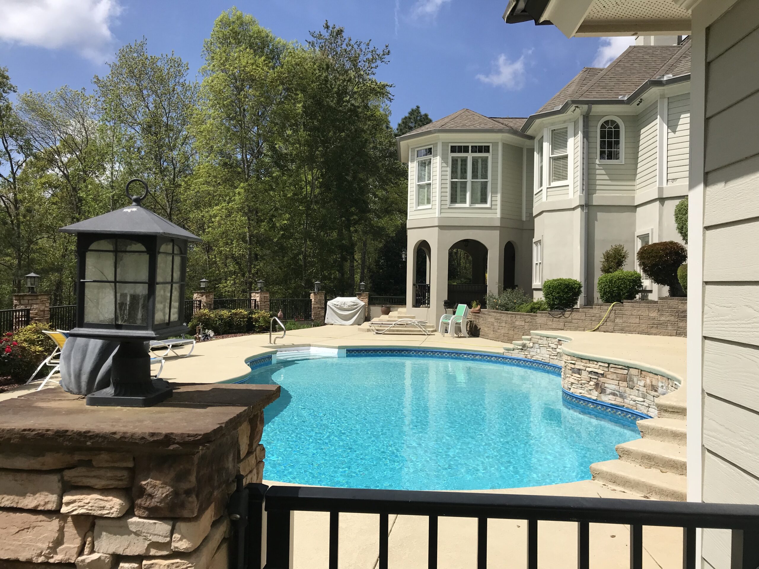 A backyard with a swimming pool surrounded by a stone patio and a two-story house with arched windows. Trees and lounge chairs are in the background.