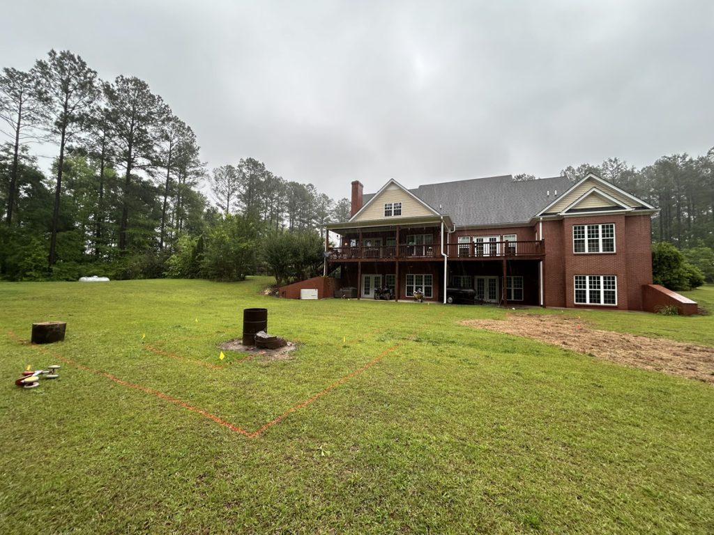 A large red-brick house with a raised deck overlooks a grassy backyard, where orange lines mark a square area near a fire pit and scattered toys. Trees border the property.