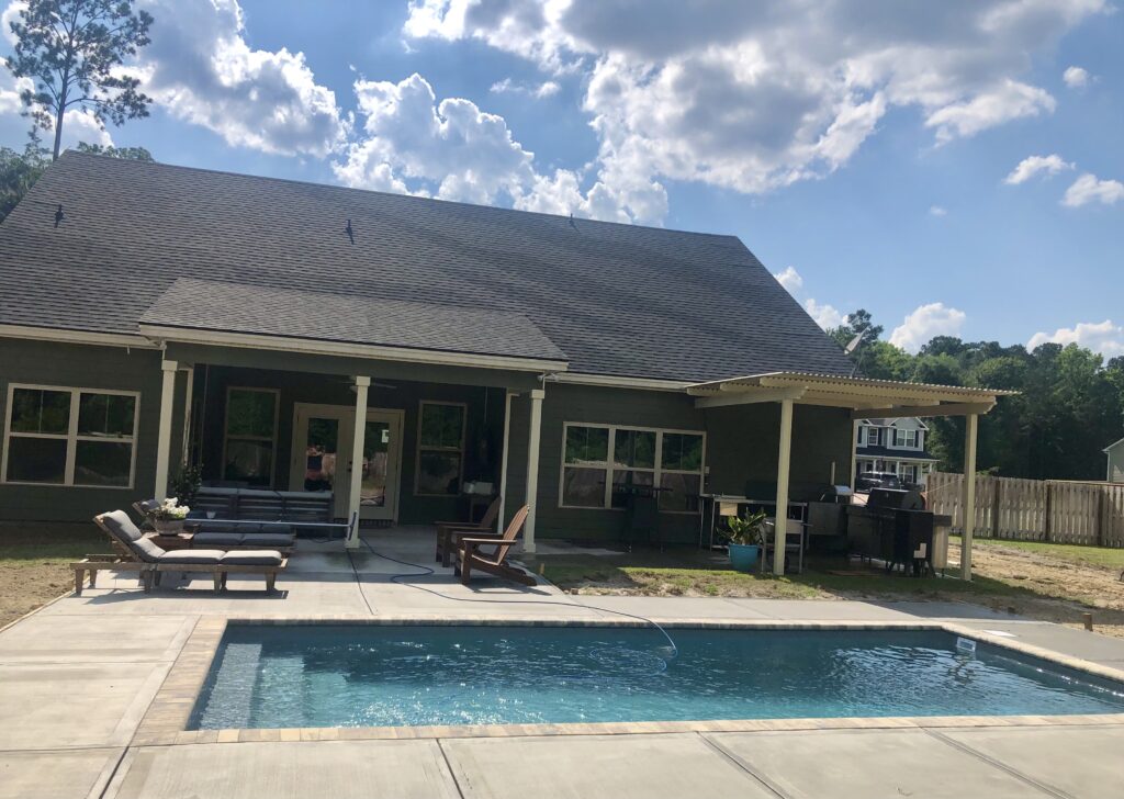 A backyard with a swimming pool, lounge chairs, covered patio, and outdoor grill under a partly cloudy sky.