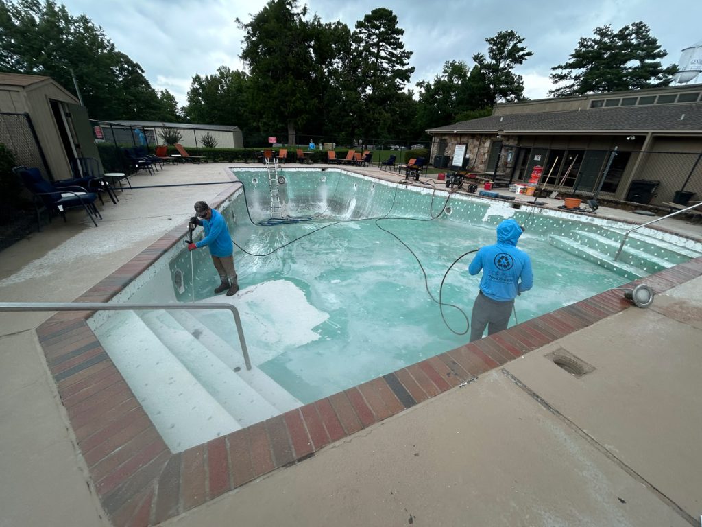 Two workers in blue shirts use power washers to clean an empty outdoor swimming pool at a facility with chairs and trees in the background.