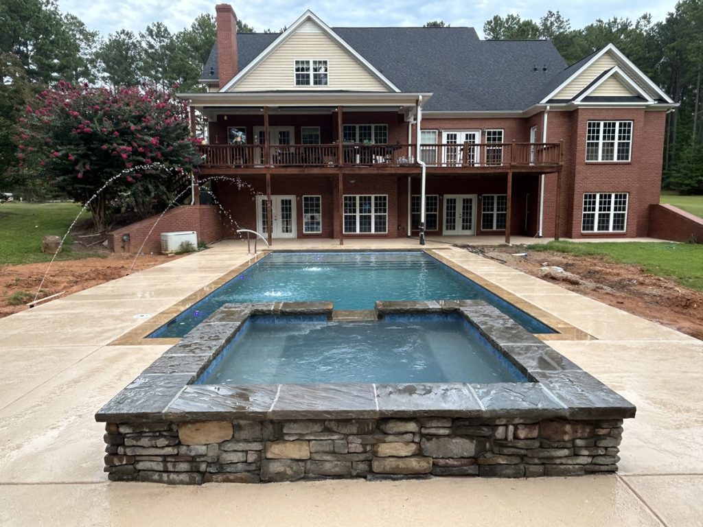 A large brick house with two stories overlooks a backyard pool and hot tub. Water jets arc into the pool, and trees and grass surround the concrete patio area.