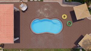 Aerial view of the Lelani model fiberglass pool surrounded by patio area, featuring lounge chairs, umbrellas, and colorful pool floats, showcasing outdoor transformation by Eco Landscaping Design.