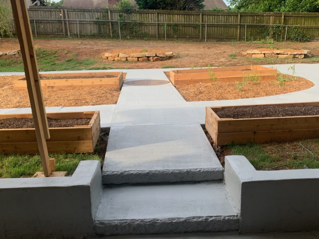 View of a backyard garden with raised wooden beds and a concrete walkway, surrounded by a fenced yard and areas of bare soil.