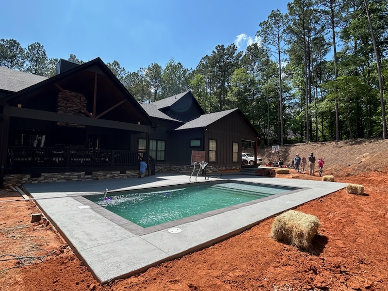 A modern house with a backyard swimming pool under shotcrete pool installation, surrounded by red soil, trees, hay bales, and several people standing near the edge of the yard.