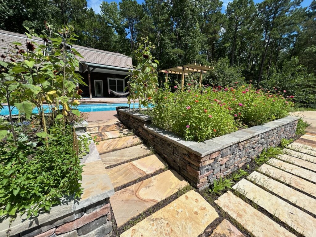 Stone pathways surround a raised flower bed and lead to a backyard pool next to a house, with trees and a pergola in the background under a sunny sky.