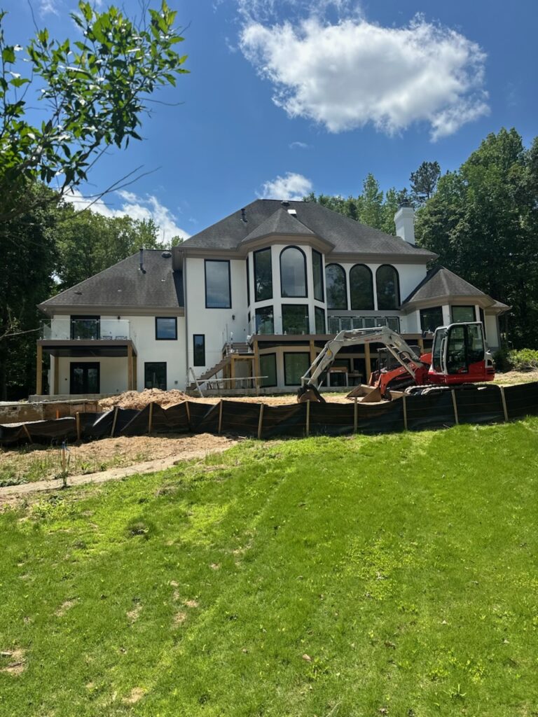A large white house with tall windows is undergoing backyard construction, with a red excavator and construction materials visible on the site.