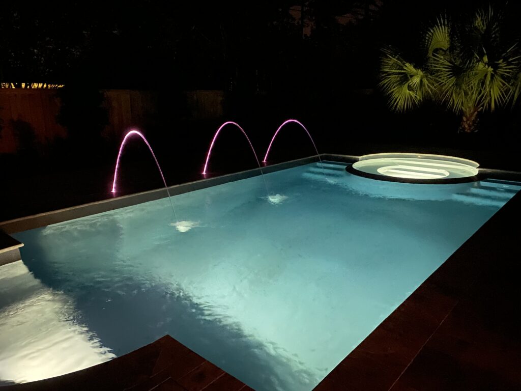 A rectangular swimming pool at night features three arcing water fountains and a circular hot tub tucked in the corner, a masterpiece crafted by expert swimming pool contractors.