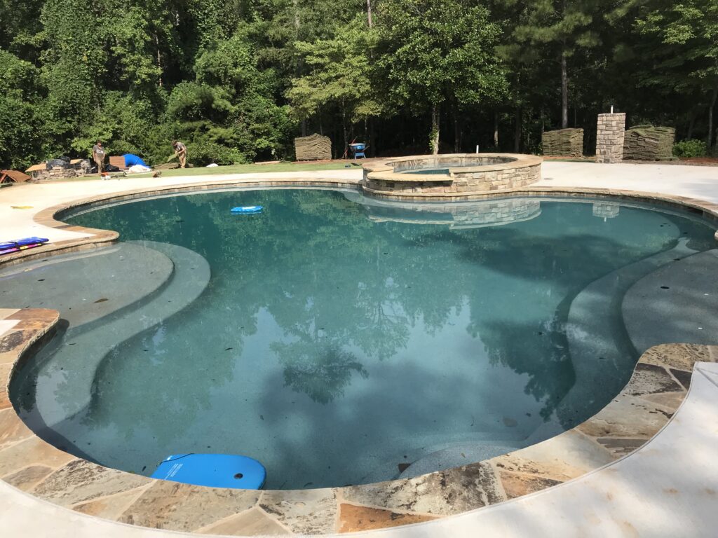 Backyard swimming pool with stone edging, surrounded by trees, offering a tranquil escape. Regular pool repairs ensure it remains a pristine oasis where nature meets relaxation.