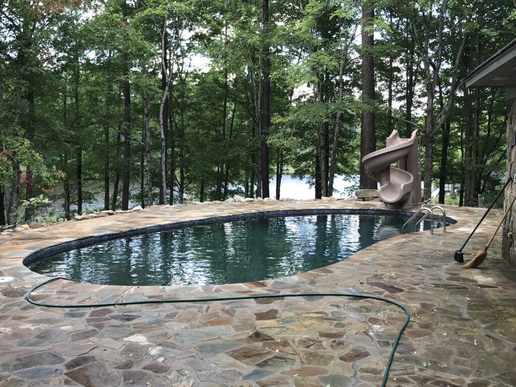 A stone patio features an inviting outdoor pool with a slide, expertly crafted by a swimming pool contractor. Surrounded by trees and overlooking a tranquil lake, the scene is complemented by a garden hose resting on the ground.