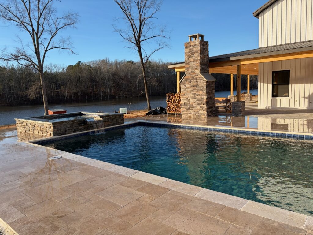A backyard pool with a hot tub, stone fireplace, and covered patio overlooks a lake and wooded area on a clear day.