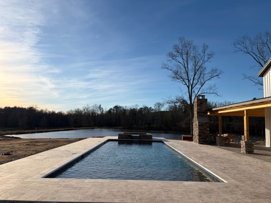 Rectangular outdoor swimming pool next to a covered patio with fireplace, overlooking a pond and trees under a clear sky at sunset.