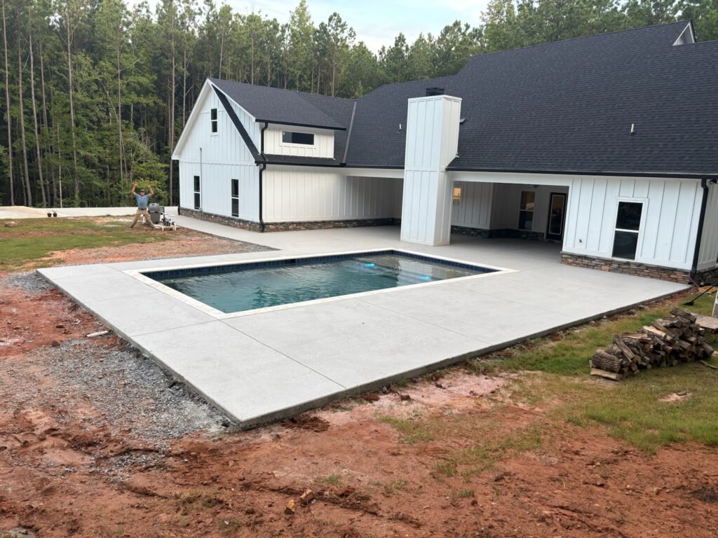 A modern white house with a dark roof features a rectangular in-ground pool surrounded by a concrete patio, with unfinished landscaping and a wooded area in the background.