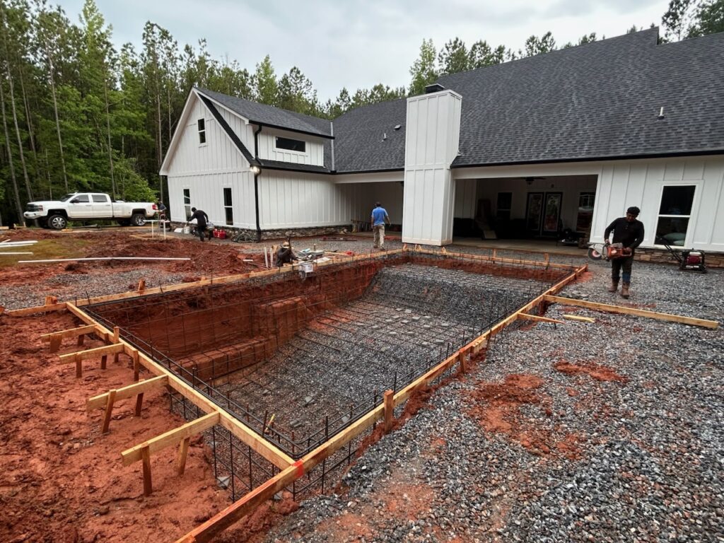 Construction workers are building an in-ground pool with rebar framework in the backyard of a white house surrounded by trees.