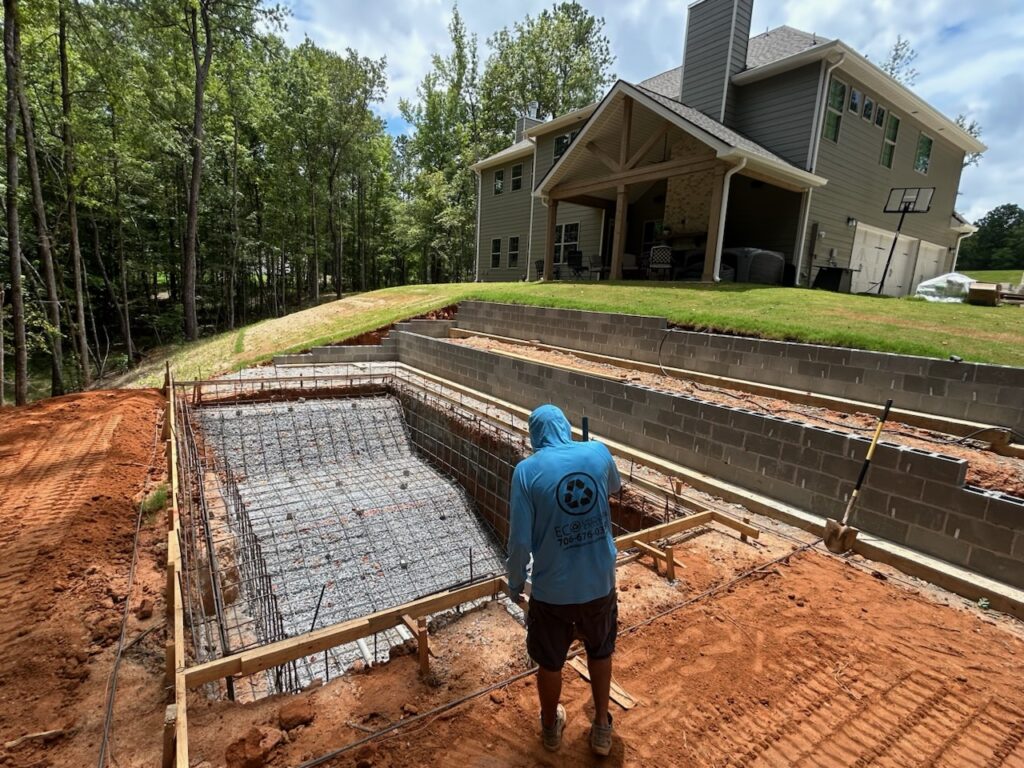 A worker stands near a rectangular in-ground pool under construction in a backyard, with a house and terraced retaining walls in the background.