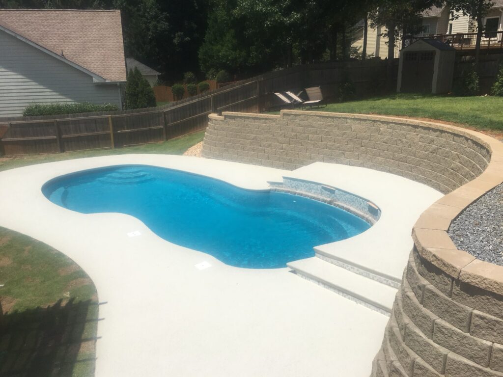 A backyard with an in-ground, kidney-shaped pool, surrounded by a concrete deck and a retaining wall, with a small lawn and wooden fencing in the background.