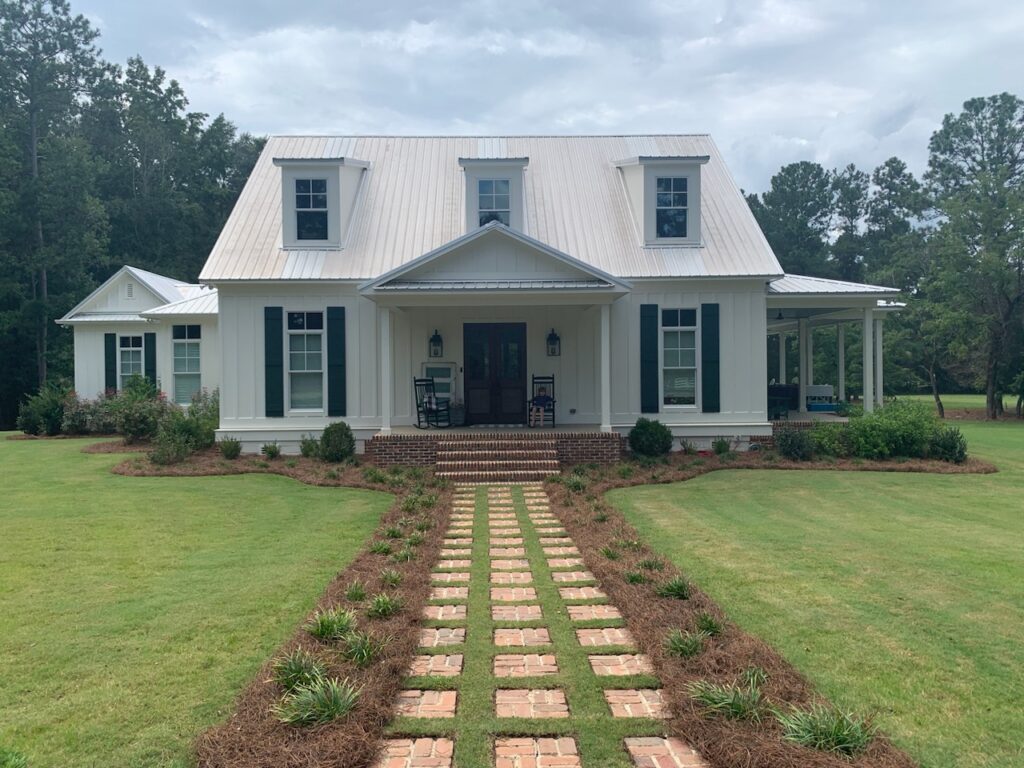 A white house with a metal roof, two dormer windows, and a covered porch. A brick path leads to the entrance, surrounded by grass and trees in the background.
