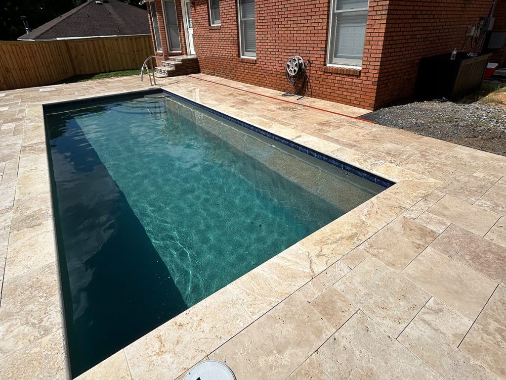 Rectangular backyard swimming pool with clear water, surrounded by stone tiles, next to a brick house with several windows and a fenced yard.
