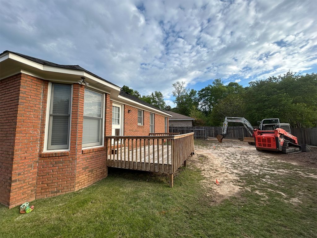 A red brick house with a small wooden deck, a grassy yard, and construction equipment on a dirt area near a wooden fence under a partly cloudy sky.