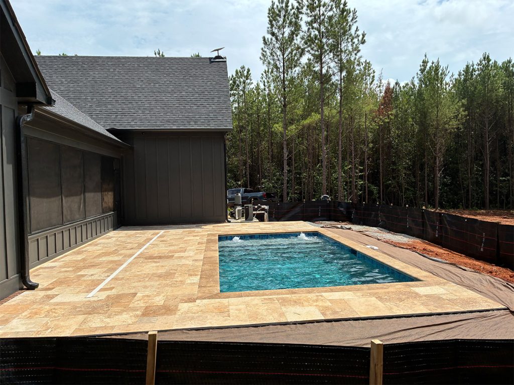 A rectangular swimming pool with tan tile decking beside a house, bordered by a black silt fence and surrounded by trees and soil under a partly cloudy sky.