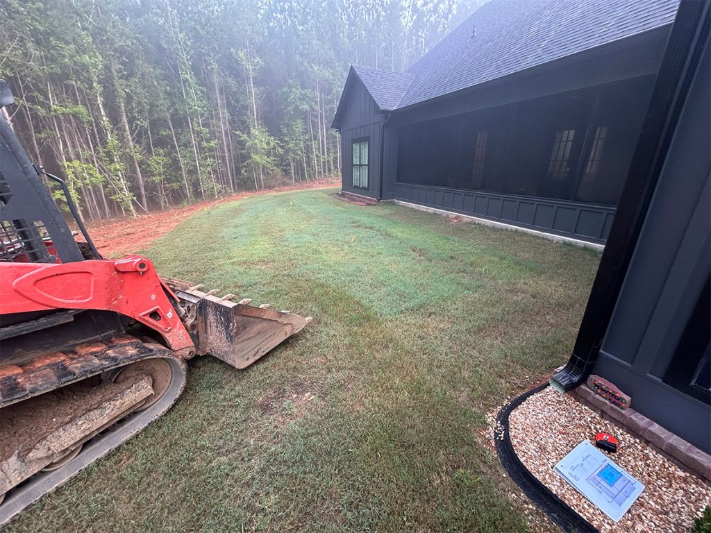 A red skid steer with a bucket is parked on freshly laid sod beside a dark-colored house with screened porch, near a wooded area.