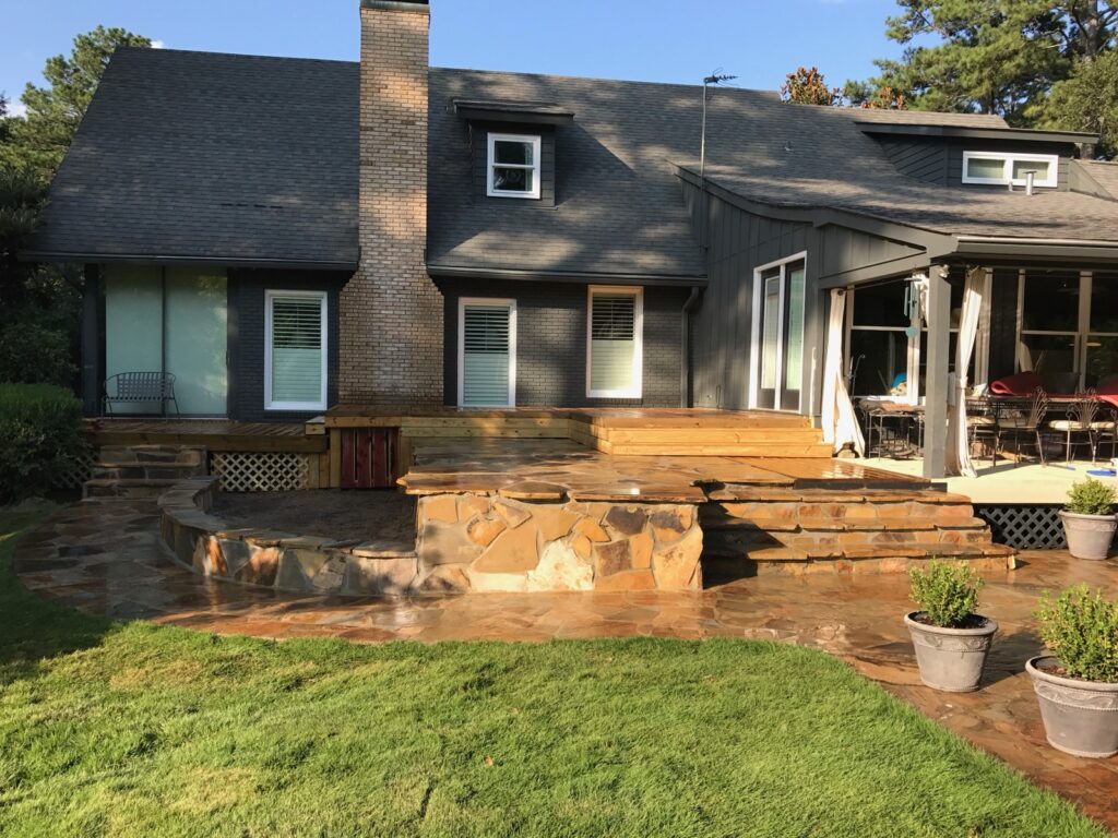 A house with gray siding and stone accents features a chimney, a multi-level wooden deck, and a stone patio area with potted plants and outdoor furniture.